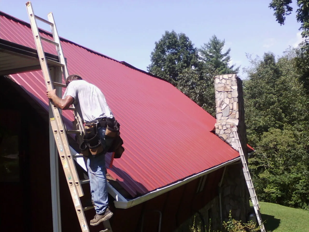 Roofer on a ladder beside a red metal roof and stone chimney — Soffit & Fascia Repair by Roofing Brothers in Cedar Creek